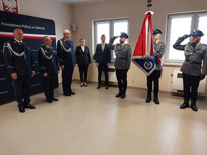 Zdjęcie w kolorze przedstawia policjantów wraz z przedstawicielami instytucji  na tle granatowej ściany z płótna z białym napisem Komenda Powiatowa Policji w Oleśnie   podczas rozpoczęcia uroczystości, po prawej poczet sztandarowy.