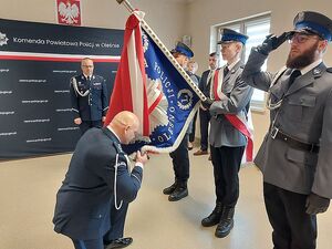 Zdjęcie w kolorze przedstawia Policjanta w umundurowaniu wyjściowym w klęku na jednej nodze przytrzymującego fragment sztandaru jako symbol objęcia stanowiska. Policjanci trzymają prawe dłonie uniesione przy czapkach.