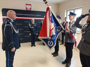 Zdjęcie w kolorze przedstawia Policjanta w umundurowaniu wyjściowym w postawie na baczność twarzą do sztandaru, policjanci trzymają prawe dłonie uniesione przy czapkach.