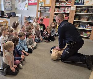 Zdjęcie w kolorze przedstawia przedszkolaki w grupie siedzą na podłodze patrzą na policjanta klęczącego nad manekinem do nauki udzielania pierwszej pomocy
