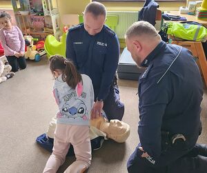 Zdjęcie w kolorze przedstawia policjantów  klęczących nad manekinem do nauki udzielania pierwszej pomocy instruując dziewczynkę.
