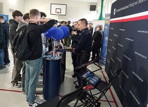 Zdjęcie w kolorze przedstawia policjantów rozmawiających z uczestnikami targów pracy na tle  ściany z płótna z napisem Komenda Powiatowa Policji w Oleśnie.
