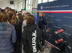 Zdjęcie w kolorze przedstawia policjantów rozmawiających z uczestnikami targów pracy na tle  ściany z płótna z napisem Komenda Powiatowa Policji w Oleśnie.