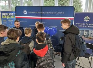 Zdjęcie w kolorze przedstawia policjantów rozmawiających z uczestnikami targów pracy na tle  ściany z płótna z napisem Komenda Powiatowa Policji w Oleśnie.