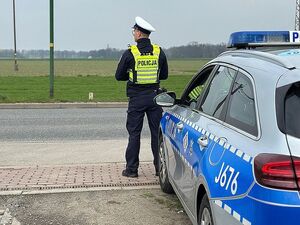 Zdjęcie w kolorze przedstawia policjanta ruchu drogowego, który stoi przed radiowozem skierowanym prostopadle do jezdni. Policjant spogląda w lewą stronę.