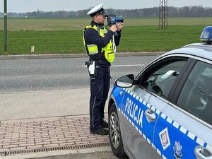 Zdjęcie w kolorze przedstawia policjanta ruchu drogowego, który stoi na krawędzi jezdni trzymając w dłoniach urządzenie pomiarowe.