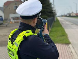 Zdjęcie w kolorze przedstawia policjanta patrzącego przez urządzenie pomiarowe w stronę jezdni.