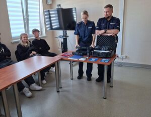 Zdjęcie w kolorze przedstawia uczniów liceum podczas zajęć z policjantami, którzy stoją przy stole na którym znajdują się materiały promocyjne oraz urządzenia do kontroli w ruchu drogowym.
