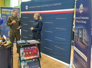 Zdjęcie w kolorze przedstawia policjantkę na stoisku promocyjnym podczas rozmowy z mężczyzna w mundurze wojskowym.