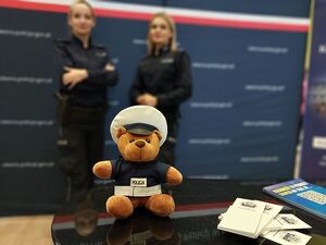 Zdjęcie w kolorze przedstawia policyjne stoisko promocyjne, zbliżenie na maskotkę misia w policyjnym mundurku w tle lekko rozmazane postaci dwóch policjantek na neibieskim tle.