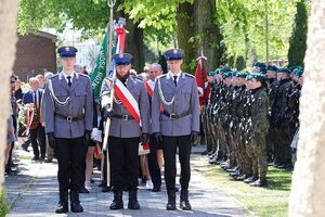 Zdjęcie w kolorze przedstawia policyjny poczet sztandarowy stojący przed pomnikiem lotników polskich.