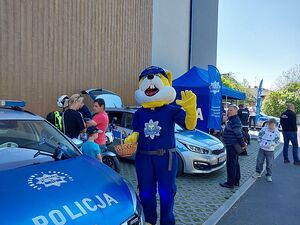 Zdjęcie w kolorze przedstawia na pierwszym planie po lewej stronie osobę ubrana w kostium -  maskotkę opolskiej policji - królika policusia tuż za nim stoisko policyjne i odwiedzający wydarzenie uczestnicy.