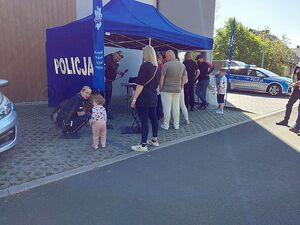 Zdjęcie w kolorze przedstawia stoisko policyjne i odwiedzających wydarzenie uczestników po lewej stronie policjantka kucająca obok małej dziewczynki, tuz za nimi pies z przewodnikiem.