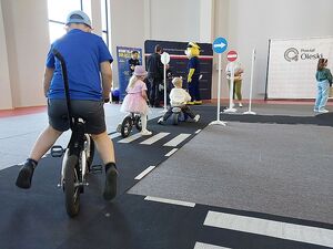 Zdjęcie w kolorze przedstawia dzieci jadące na rowerkach po miasteczku ruchu drogowego, w tle ścianka z płótna z napisem Komenda Powiatowa Policji w Oleśnie i stojący przed nią policjanci.
