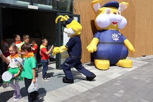 Zdjęcie w kolorze przedstawia przedszkolaków wychodzących z budynku przed którym stoi pompowana maskotka policusia oraz osoba przebrana w policusia