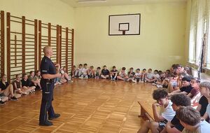 Zdjęcie w kolorze przedstawia policjanta stojącego na środku sali gimnastycznej do koła siedzą dzieci.