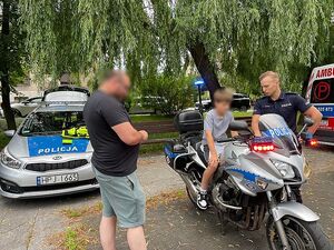 Zdjęcie w kolorze przedstawia dziecko siedzące na motocyklu policyjnym, radiowóz policyjny oraz policjanta