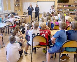 Zdjęcie w kolorze przedstawia dzieci siedzące w szkolnych ławkach, policjantki stoją przy tablicy.