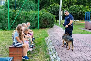 Zdjecie w kolorze przedstawia dzieci siedzące na ławce na terenie zielonym, przed nimi stoi policjant w psem. W tle drzewa.