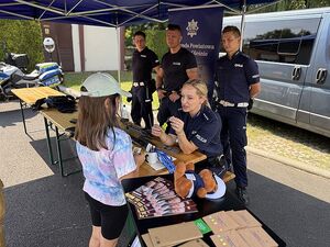 Zdjecie w kolorze przedstawia policjantkę wręczająca dziewczynce element odblaskowy w tle stoją policjanci.
