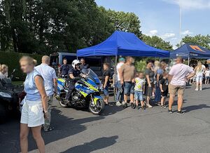 Zdjęcie w kolorze przedstawia plac zewnętrzny KPPSP w Oleśnie na którym prezentowane są pojazdy oraz sprzęt służb - Policji i Straży Pożarnej przy których stoją odwiedzający - dorośli wraz z dziećmi.