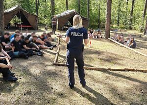 Zdjęcie w kolorze przedstawia po środku policjantkę dalej młodzież siedzącą na ściółce leśnej pośród drzew,  w tle namioty i las.