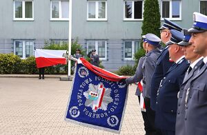 Zdjęcie w kolorze przedstawia policjantów w mundurach galowych prezentujących sztandar  podczas uroczystych obchodów Święta Policji w Oleśnie. W tle Poczet Flagowy wciągający biało czerwoną flagę na maszt.