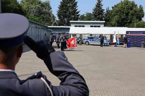 Zdjęcie w kolorze przedstawia policjanta oddającego honory w tle Poczet Sztandarowy i stojący za nim policjanci.