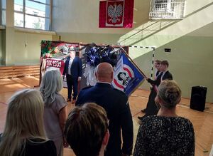 Zdjęcie w kolorze przedstawia inaugurację roku szkolnego, na sali gimnastycznej wstępuje poczet sztandarowy szkoły.