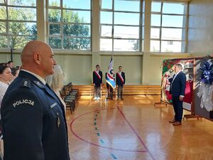 Zdjęcie w kolorze przedstawia inaugurację roku szkolnego, na sali gimnastycznej na pierwszym planie po lewej policjant w umundurowaniu galowym po środku poczet sztandarowy szkoły.