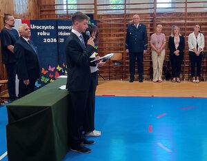 Zdjęcie w kolorze przedstawia inaugurację roku szkolnego, na sali gimnastycznej po lewej stronie dyrektor oraz prowadzący a na środku policjant w mundurze galowym oraz inni goście.
