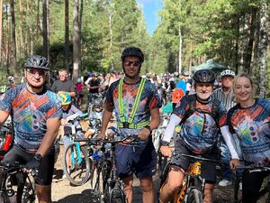 Zdjęcie w kolorze przedstawia policjantów w strojach sportowych stojących jeden obok drugiego z rowerami przy boku, w tle pozostali uczestnicy wydarzenia i las.