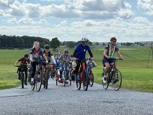 Zdjęcie w kolorze przedstawia peleton jadący drogą wśród łąk, w tle obłoki na błękitnym niebie.
