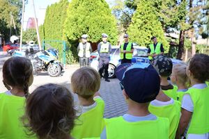 zdjęcie przedstawia przedszkolaki w kamizelkach odblaskowych spoglądajace na policjantów na placu przy przedszkolu
