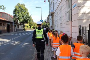 zdjęcie przedstawia dzieci ubrane w kamizelki odblaskowe idące chodnikiem , obok nich idzie policjant.