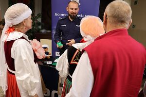 Zdjęcie w kolorze przedstawia seniorów w strojach ludowych przed stoiskiem policyjnym