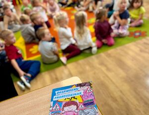 Zdjęcie przedstawia książkę " bezpieczny przedszkolak" na blacie ławki szkolnej w tle grupę dzieci siedzących na podłodze.