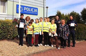 Zdjęcie przedstawia policjantów wraz z dziećmi w kamizelkach odblaskowych, którzy pozują do wspólnego zdjęcia na tle budynku z napisem Policja.