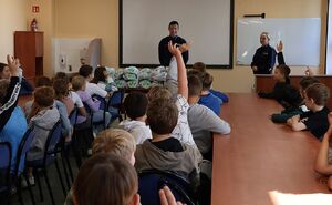Zdjęcie przedstawia policjanta i policjantkę wręczających kaski dla dzieci w szkole.
