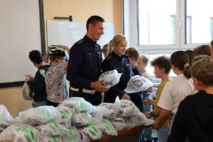 Zdjęcie przedstawia policjanta i policjantkę wręczających kaski dla dzieci w szkole.