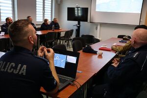 Zdjęcie przedstawia policjantów w sali odpraw, patrzą na slajd wyświetlony na ekranie ściennym.