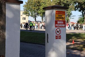 Zdjęcie przedstawia  furtkę - wejście na teren szkoły, w oddali osoby zgromadzone w miejscu ewakuacji.