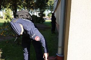 Zdjęcie przedstawia policjanta sprawdzającego teren szkoły, funkcjonariusz pochyla się nad odpływem rynny.