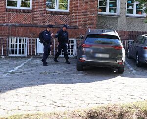 Zdjęcie przedstawia policjantów sprawdzających teren szkoły - parking  przy użyciu zwierciadła na kiju.