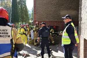 Zdjęcie przedstawia policjantów  i strażaków przy budynku z czerwonej cegły, na pierwszym planie dowódcy akcji w oznaczonych kamizelkach.