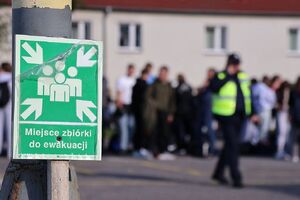 Zdjęcie przedstawia zieloną tabliczkę z napisem miejsce do ewakuacji w tle rozmazane osoby.