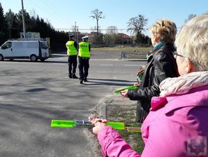 przedstawicielki stowarzyszenia nasza szansa trzymają w ręce upominki w postaci odblasków i długopisów, w tle dwaj policjanci idą w stronę jezdni, przejeżdża pojazd dostawczy