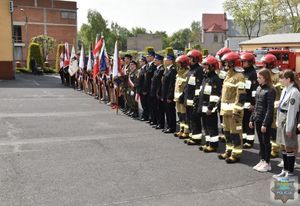 Stojący w szeregu harcerze i  strażacy  w umundurowaniu bojowym oraz wyjściowym na dalszym planie poczty sztandarowe przedstawicieli szkół średnich, straży pożarnej i policji, w tle wóz bojowy straży pożarnej.