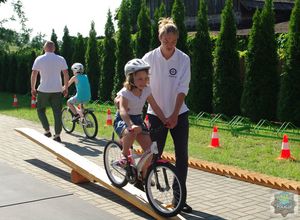 Dziewczynka na rowerze pokonuje przeszkodę. Asekuruję ją pracownik Akademii Bezpieczeństwa.