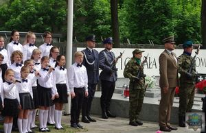 Dzieci śpiewają piosenki patriotyczne. W tle warta honorowa wystawiona przez służby mundurowe.